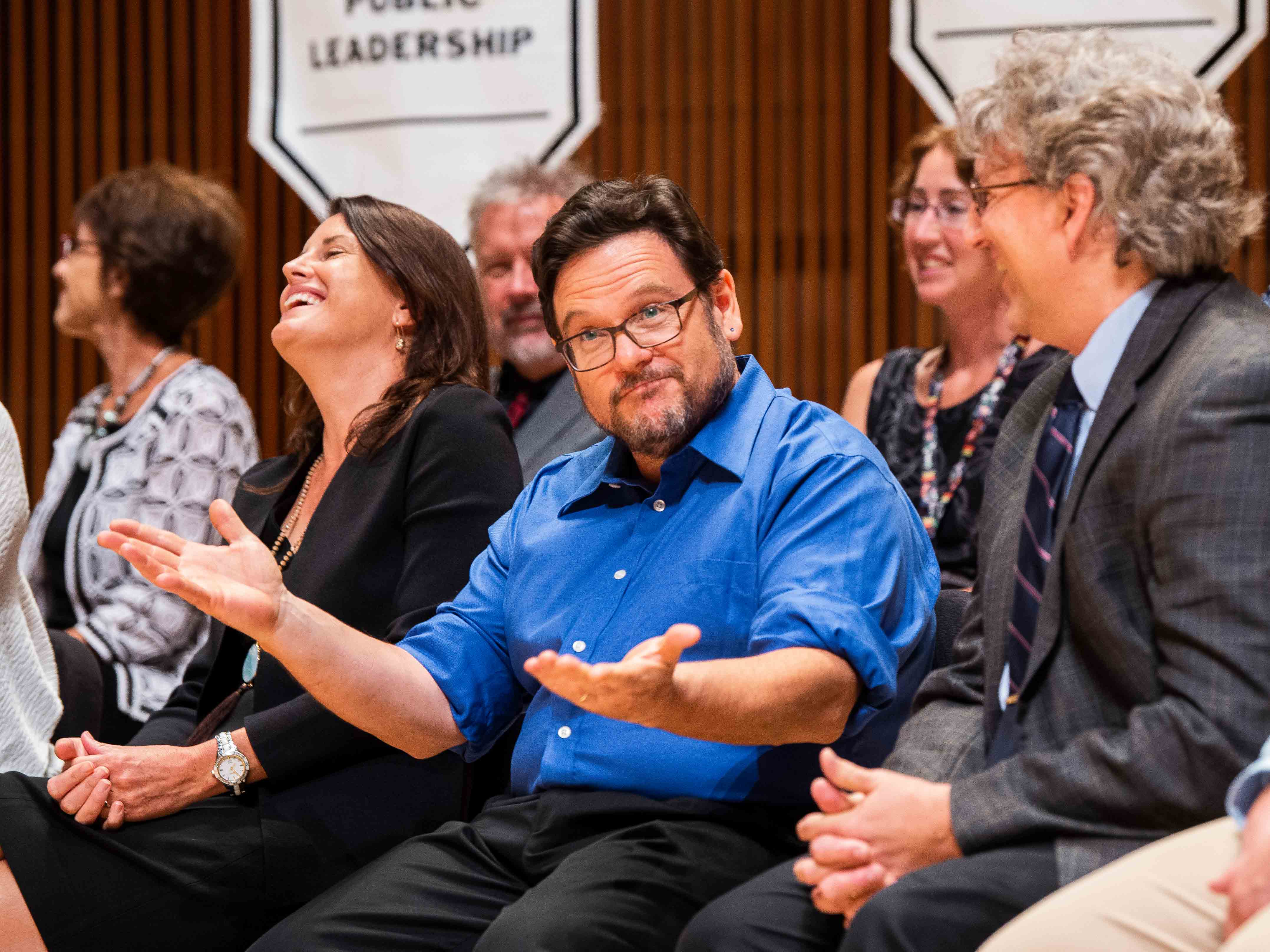 Seated bearded man in blue shirt looks off to the side with half-smile on his face while people on either side of him laugh