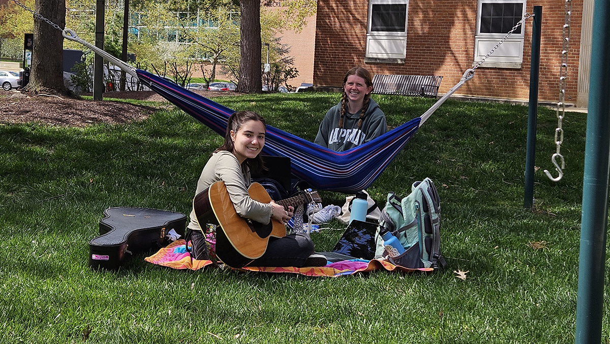 student sitting on blanket with a guitar in front of a student smiling in a hammock