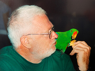White-haired man with glasses feeds cracker to a tropical bird on his shoulder