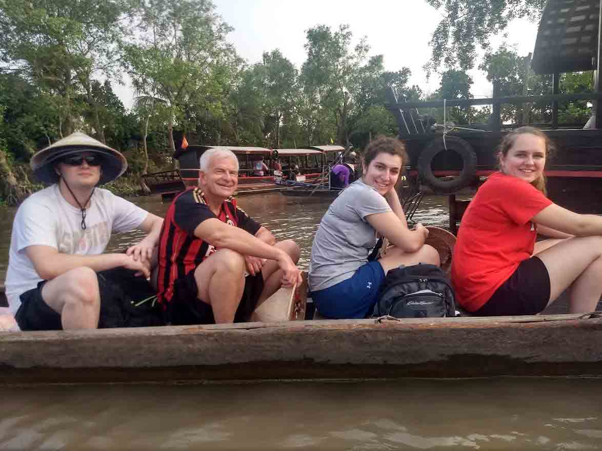 Three students sit on a wooden river boat in Vietnam with their professor