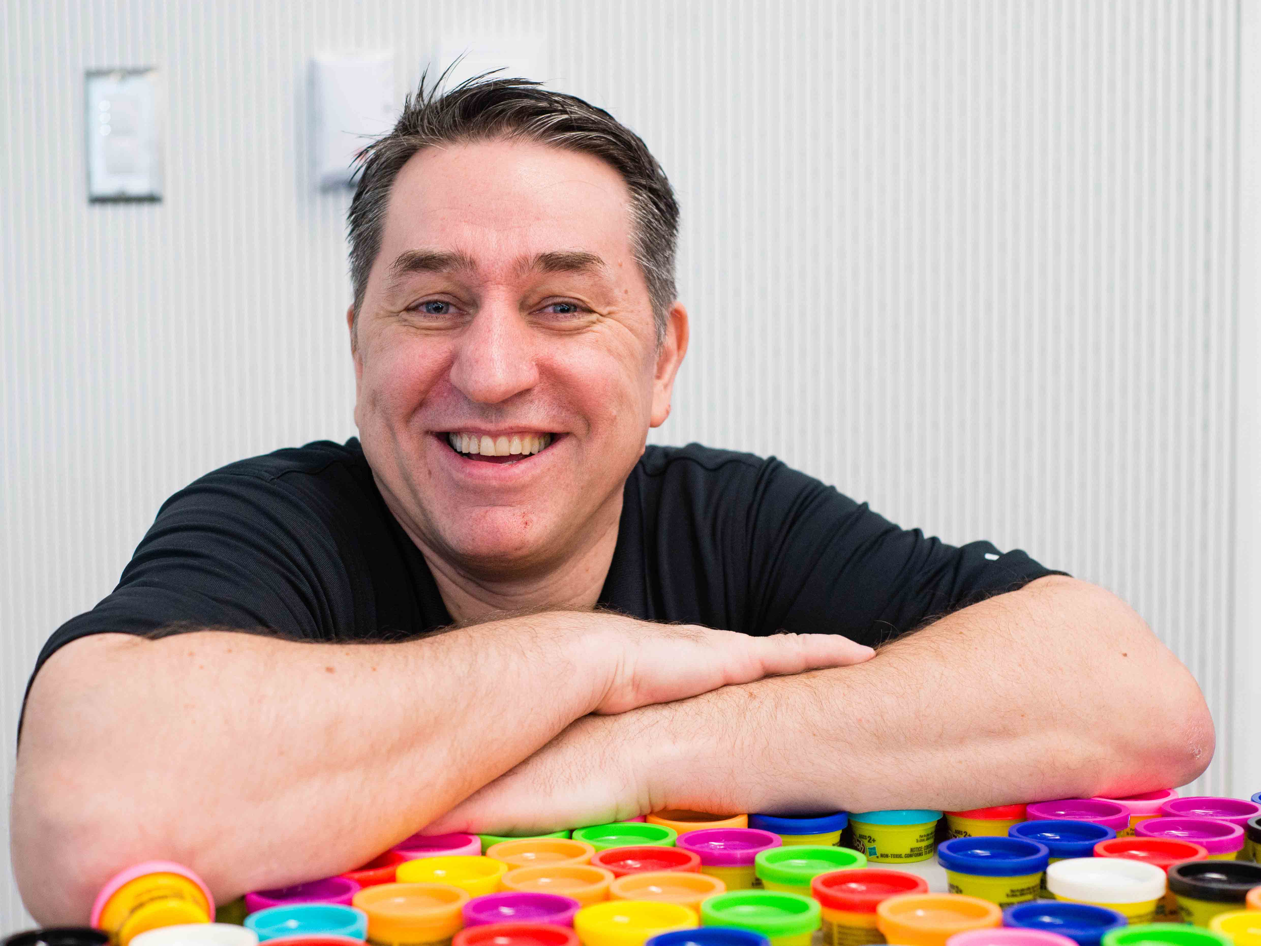 Dark-haired man smiles with his arms resting atop an array of small containers of Play-Doh