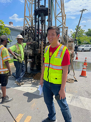 Student wearing red Scholars shirt and neon yellow safety vest stands at road construction site