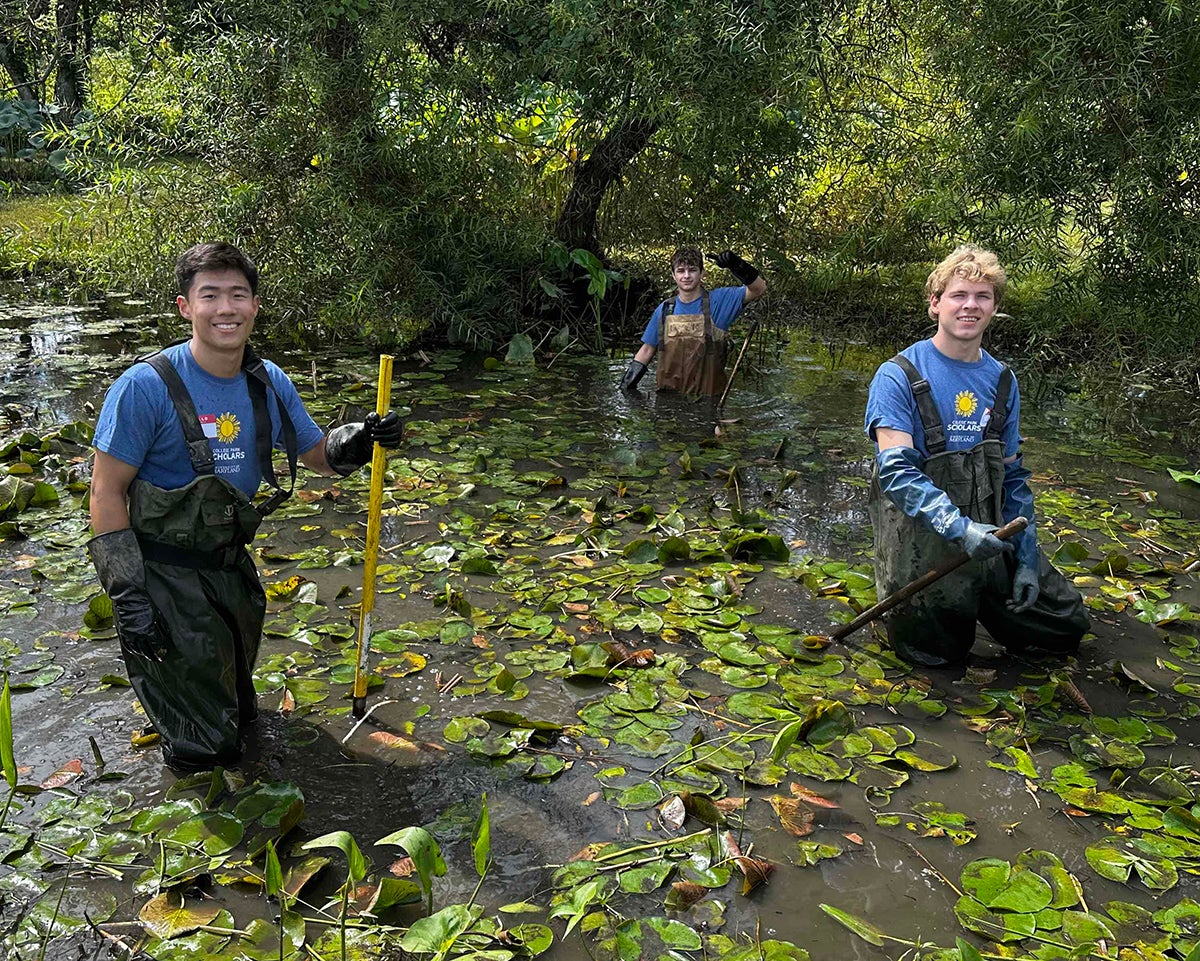 smiling male students in blue shirts and green coveralls in knee deep pond