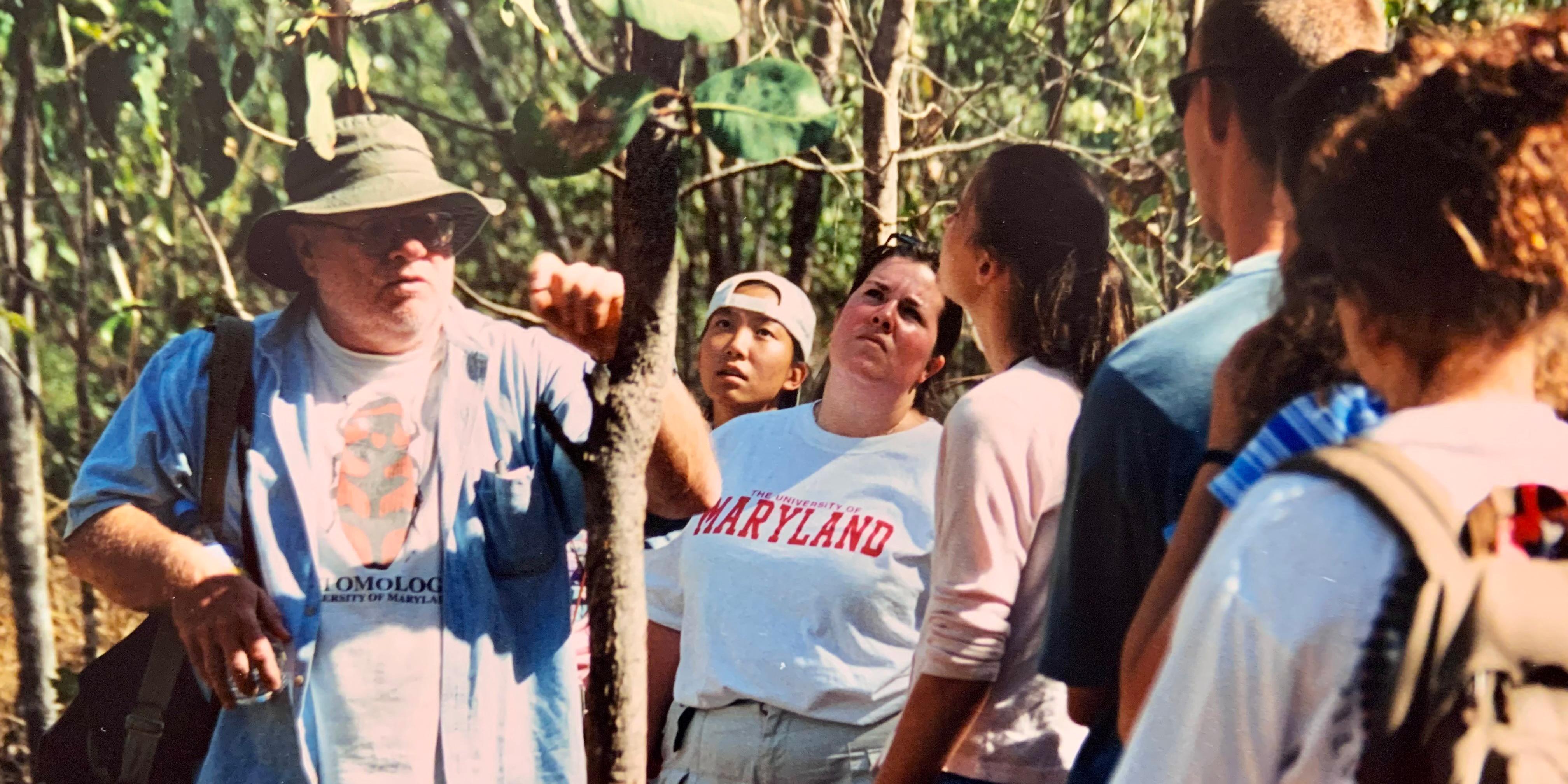 Man in hat shows off snake curled in tree in jungle as students look on