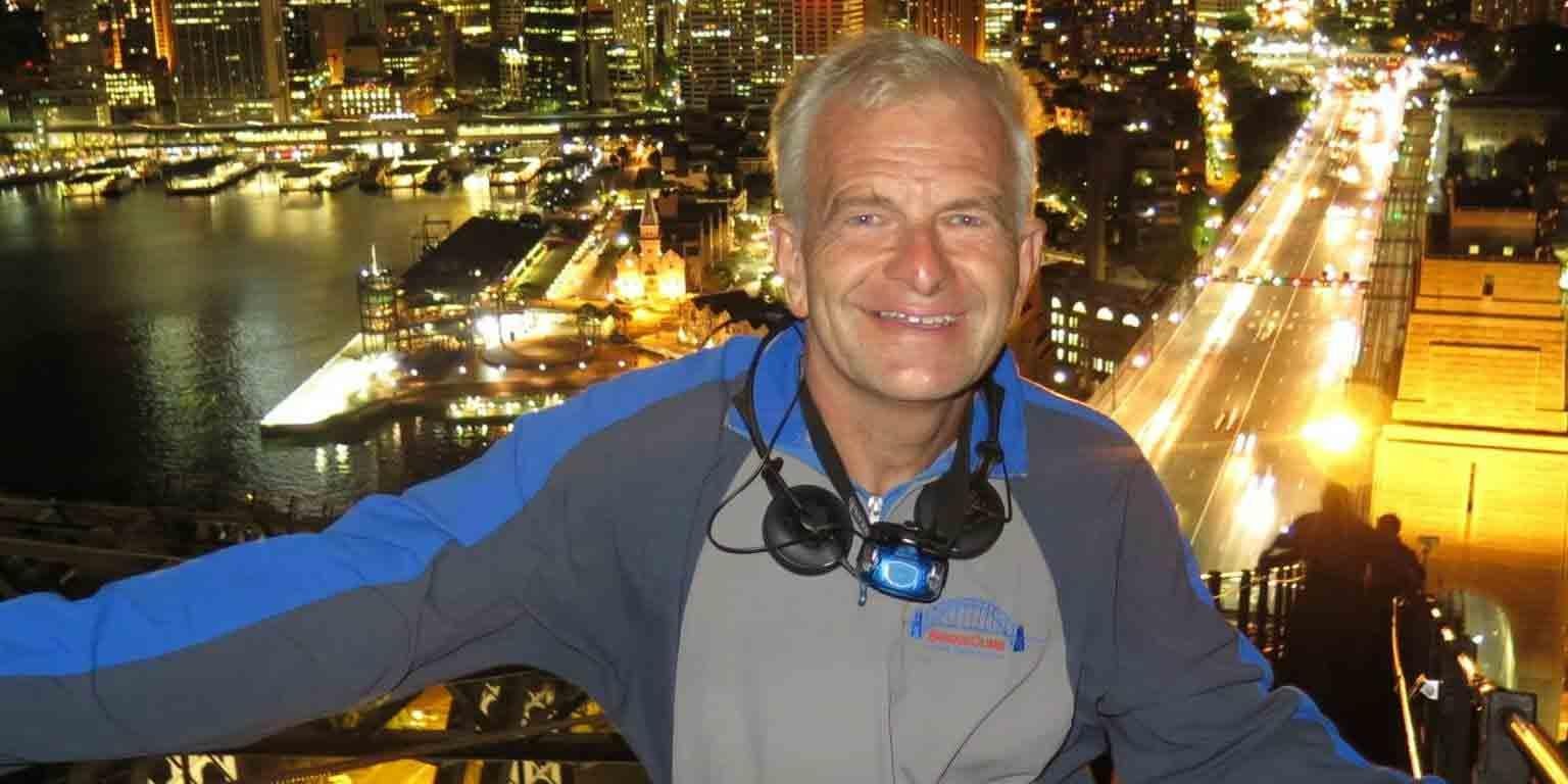 Man stands smiling near top of Sydney Bridge at night with Sydney city lights behind him