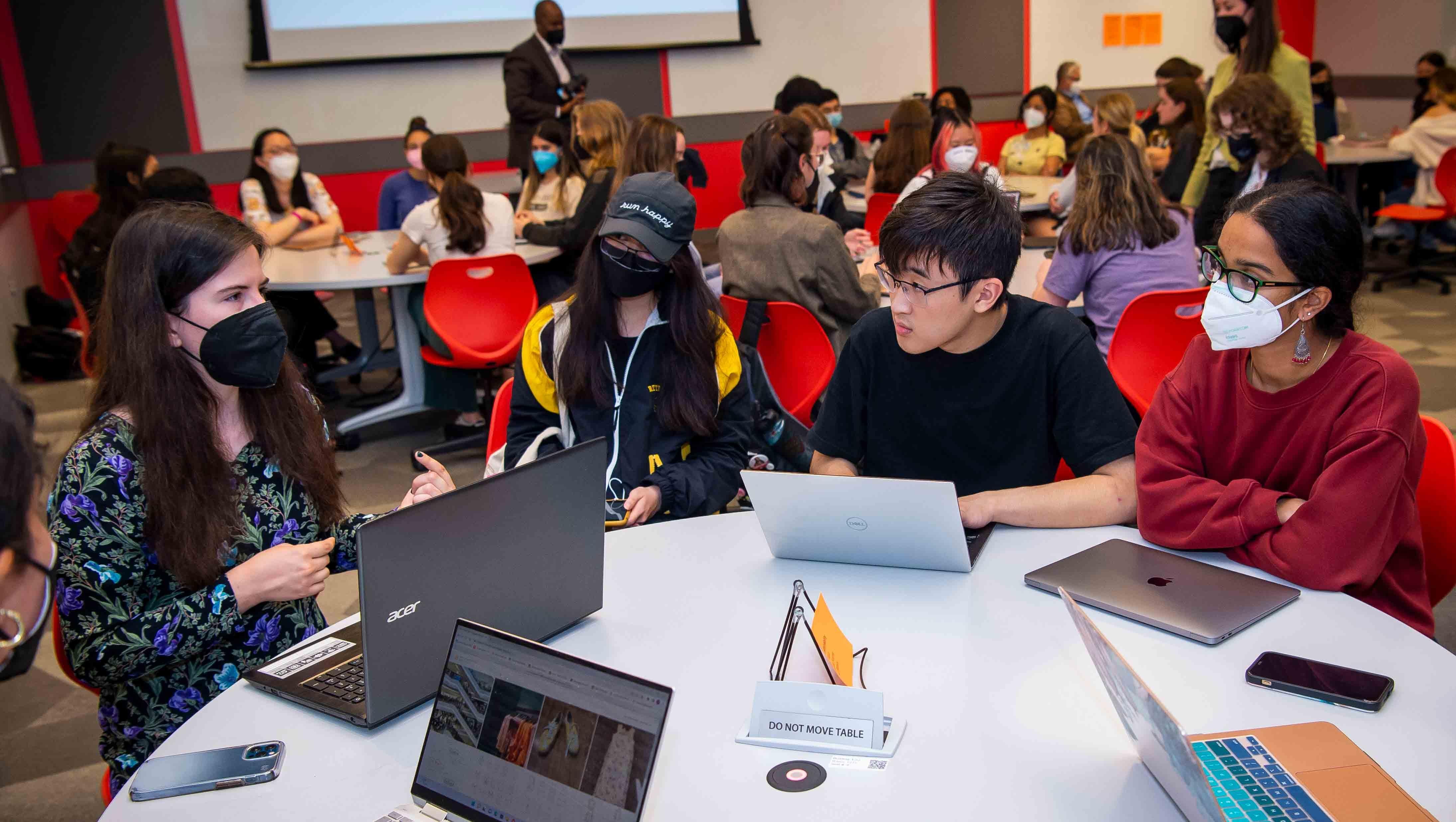 Students gathered around a table with laptops, listening to a female student talking