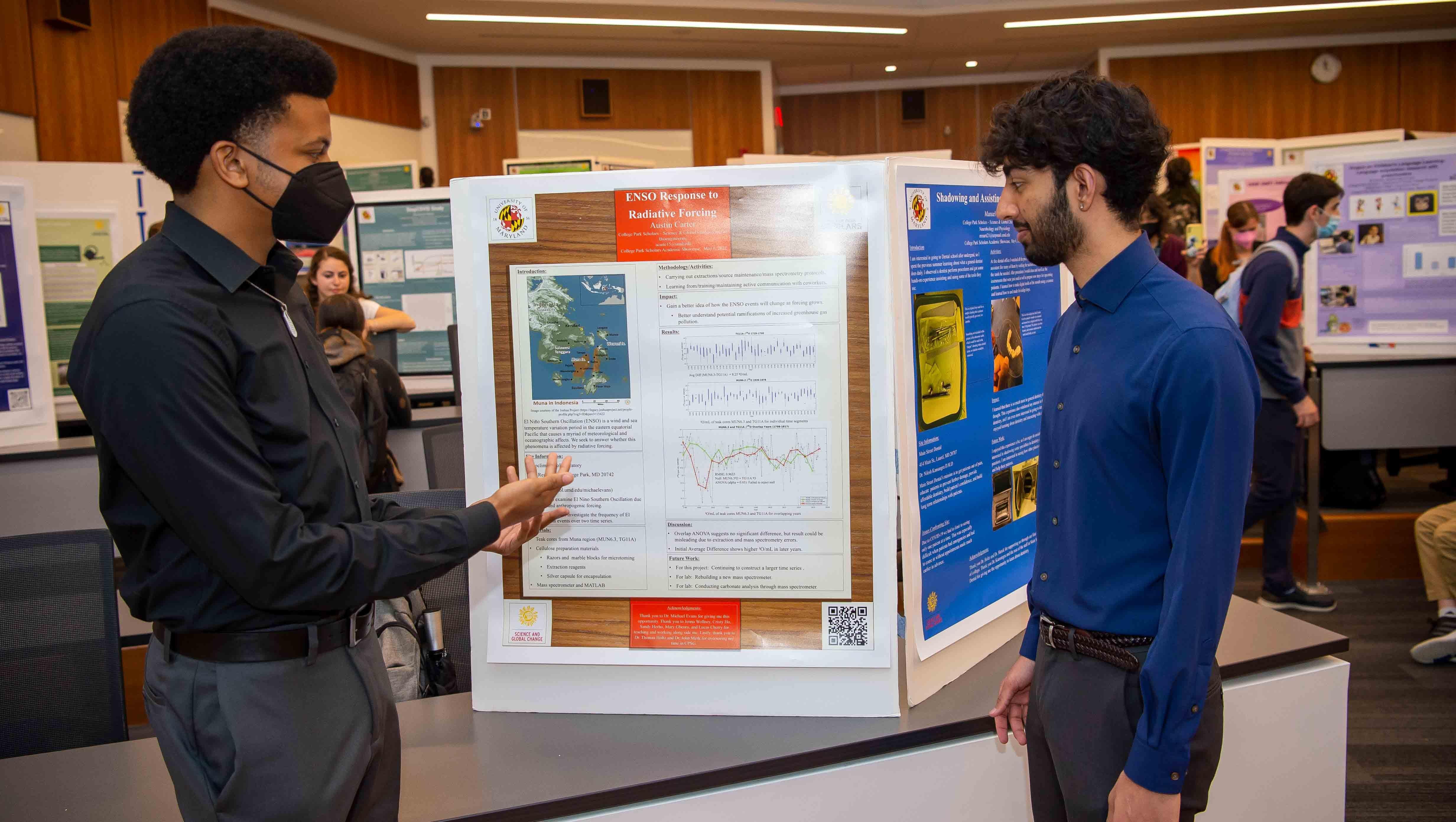 One male student gestures at an academic poster while a second male student looks on