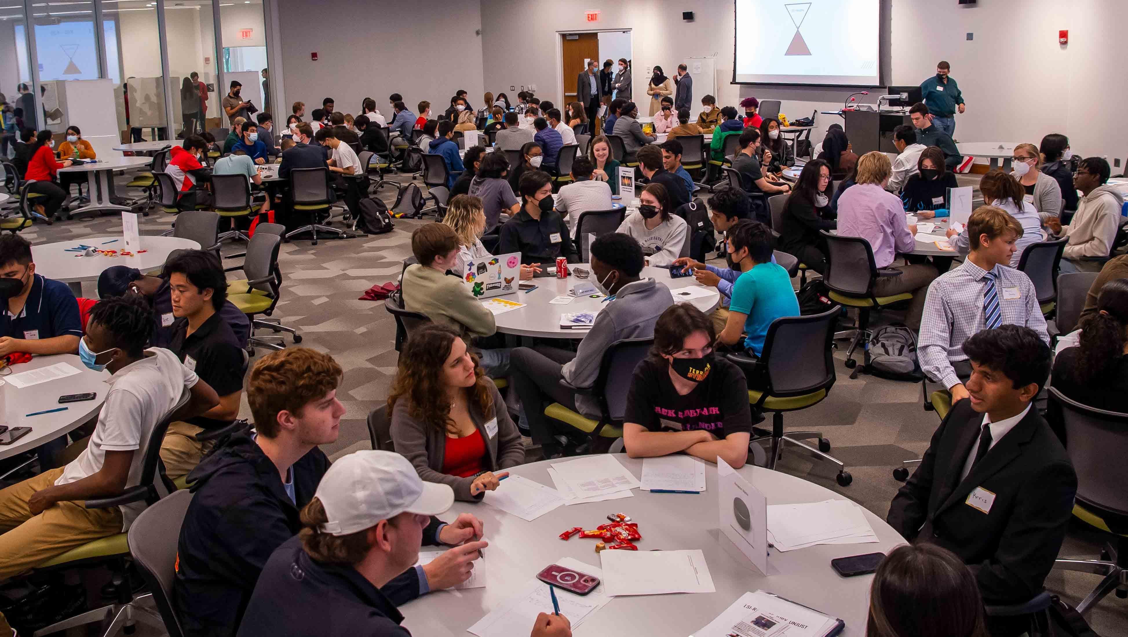 Photo shows room with several large tables with students seated around them, engaged in discussion