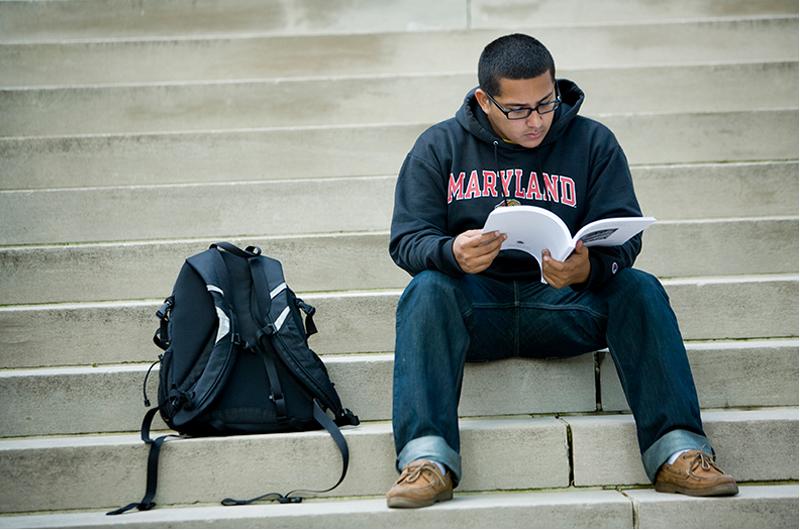student in blue sweatshirt sitting on steps with a notebook and bookbag