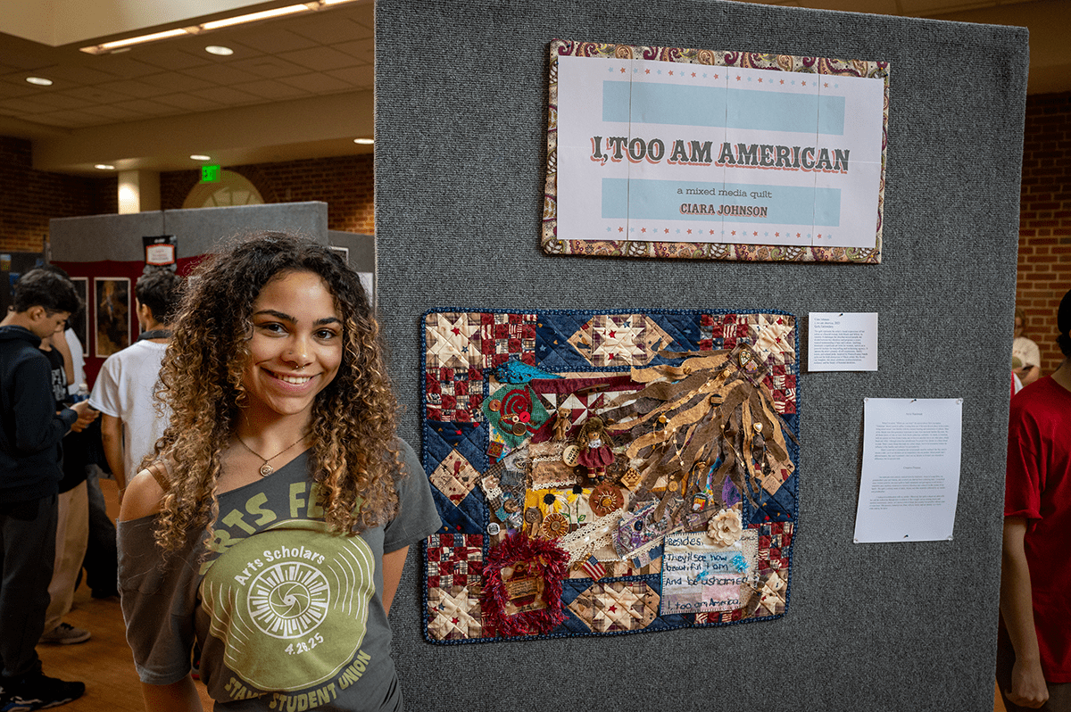 woman in gray ArtsFest t-shirt standing next to a gray display board