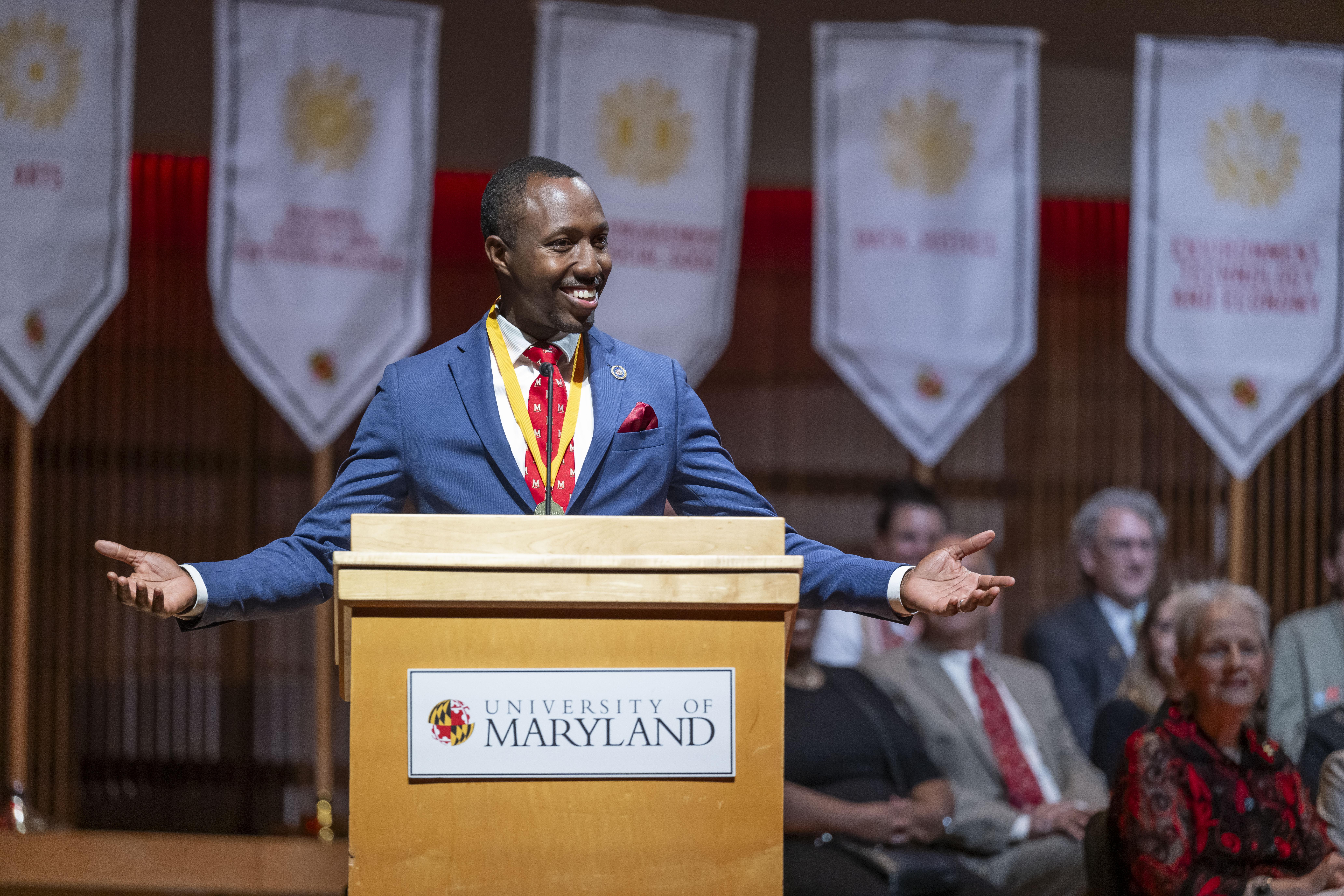 man in blue suit at podium smiling with arms outstretched
