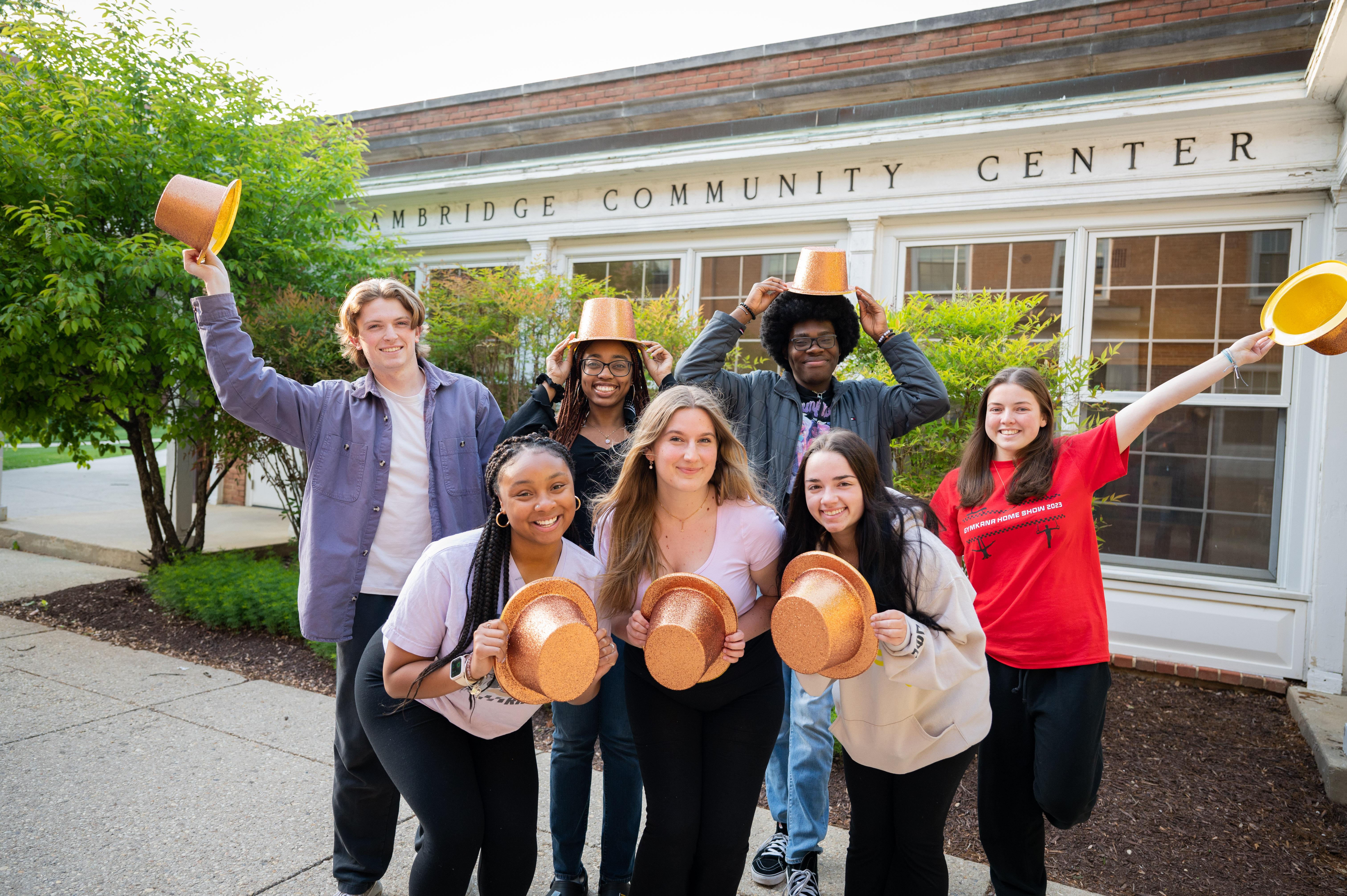 students holding gold top hats smiling in front of Cambridge Community Center sign on brick building