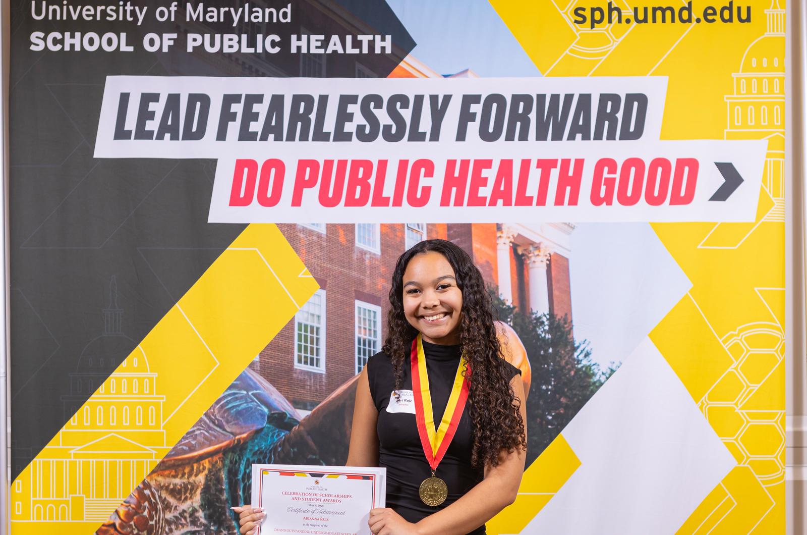 woman in black dress with a red and yellow medallion around her neck holding a certificate in front of a sign