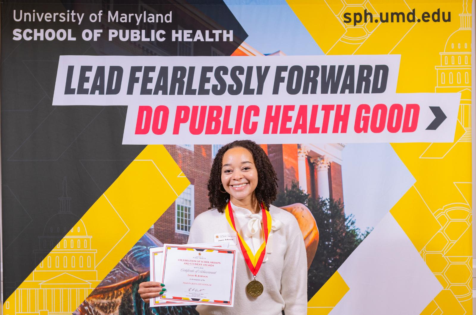 woman with a red and yellow medallion around her neck holding a certificate in front of a sign
