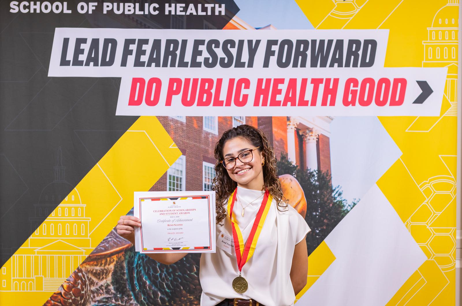 woman with a red and yellow medallion around her neck holding a certificate in front of a sign
