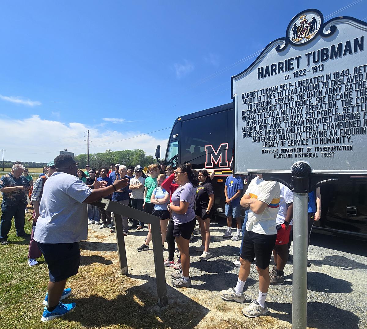 man facing students in front of a black Maryland bus, Harriet Tubman marker is in the foreground