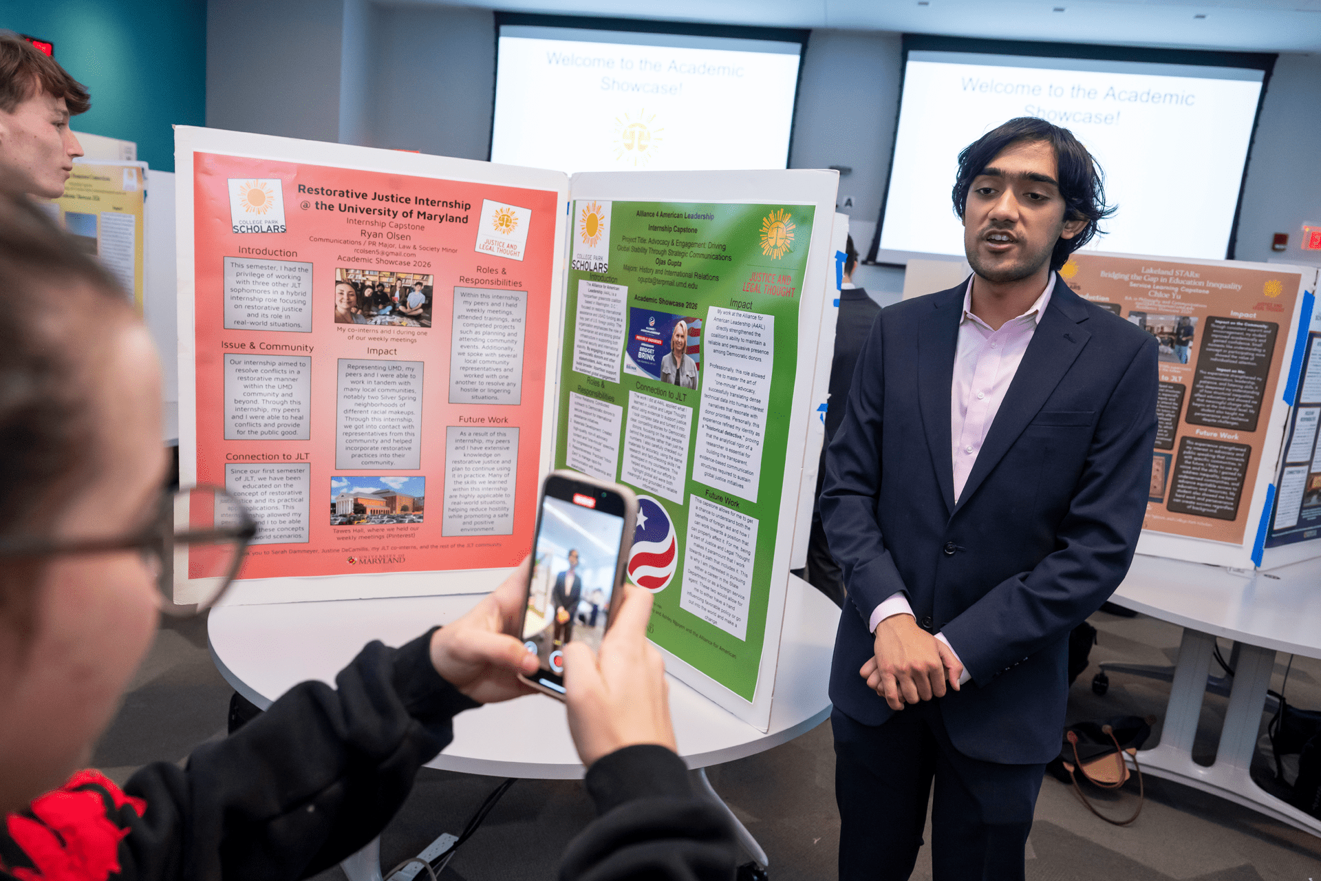 student standing next to a poster board being recorded on a phone by another student