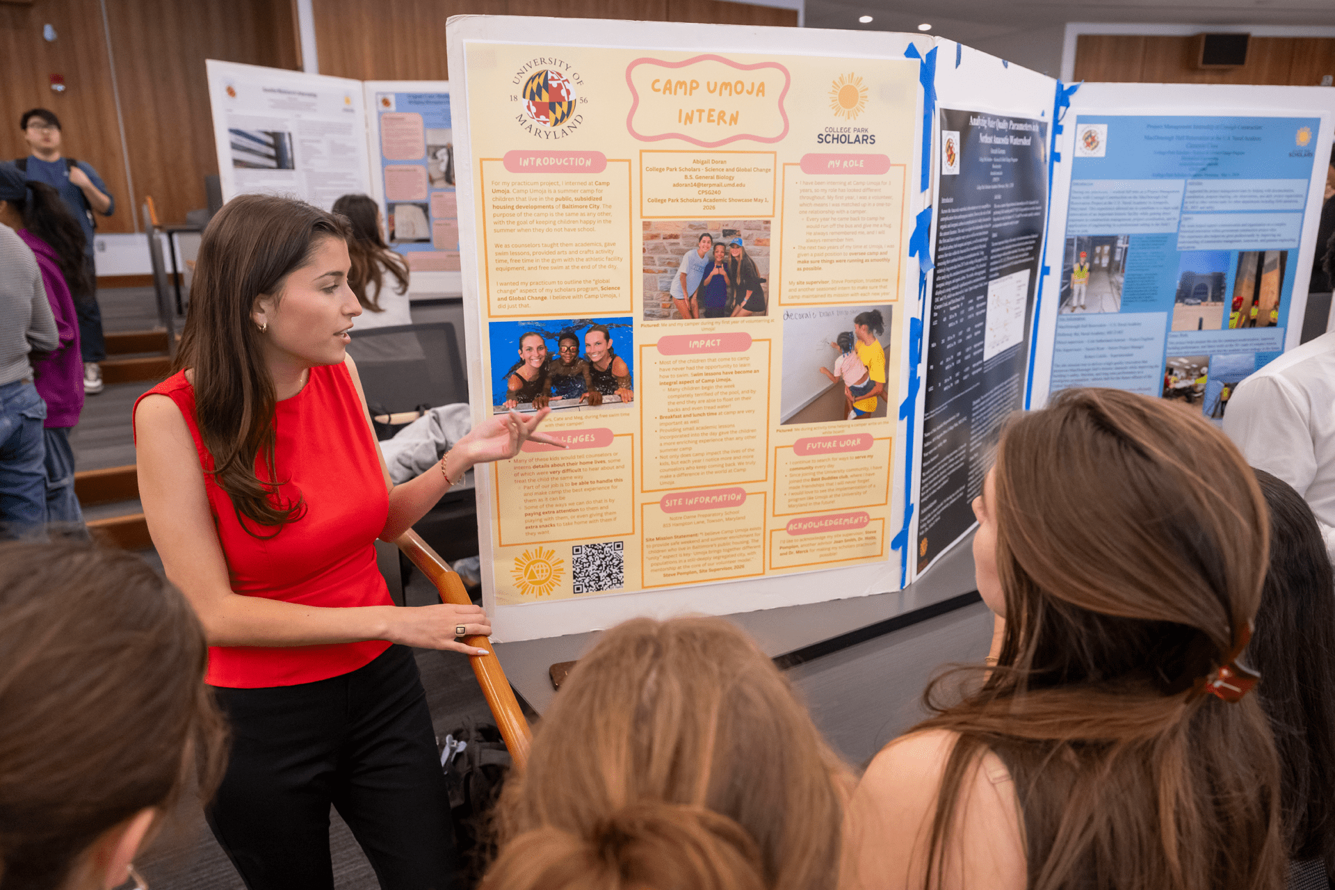woman in red shirt gesturing toward poster board in front of other students